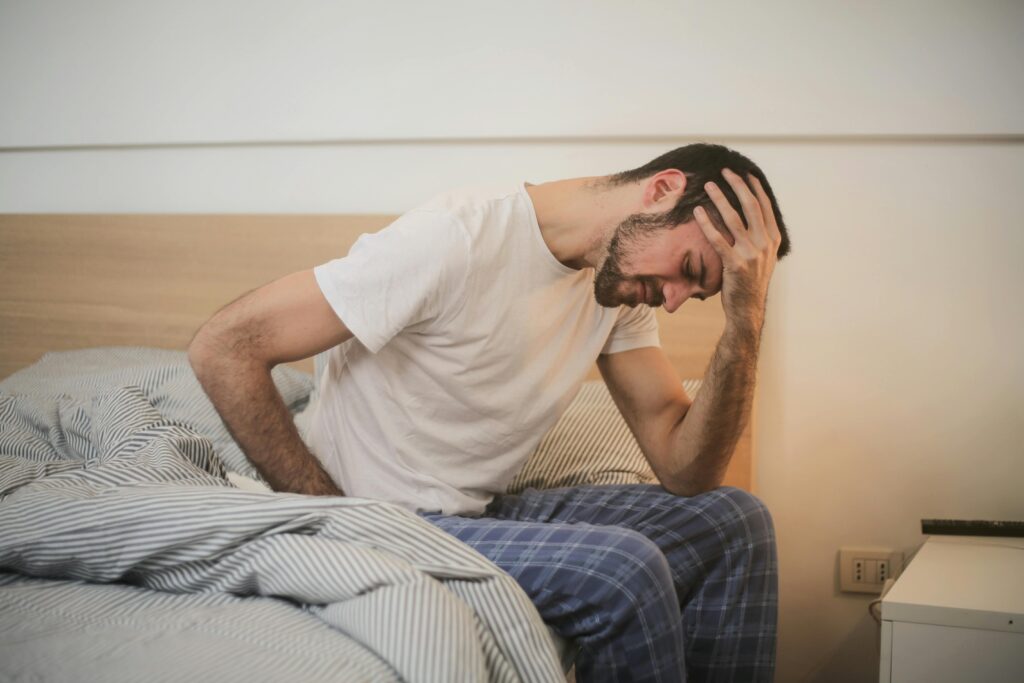 A young man in pajamas holding his head, sitting on a bed.